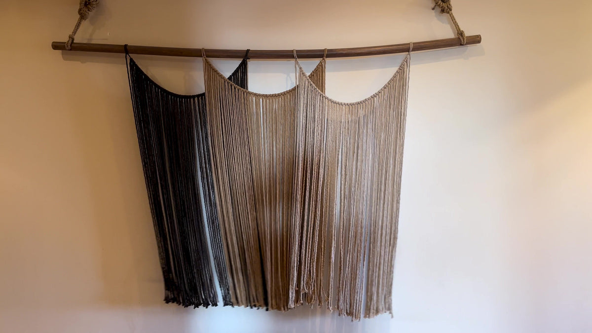 Suspenso Terra, a large handmade macramé wall hanging in charcoal, caramel and taupe by Nyra Atelier, on a warm plaster wall in the Porto atelier