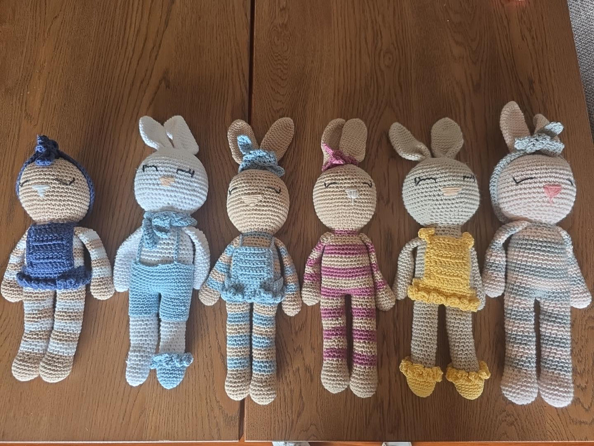 The Nyra Bunny Family by Nyra Atelier: six handmade crochet bunnies lying side by side on a wooden table, each in its own colours and outfit
