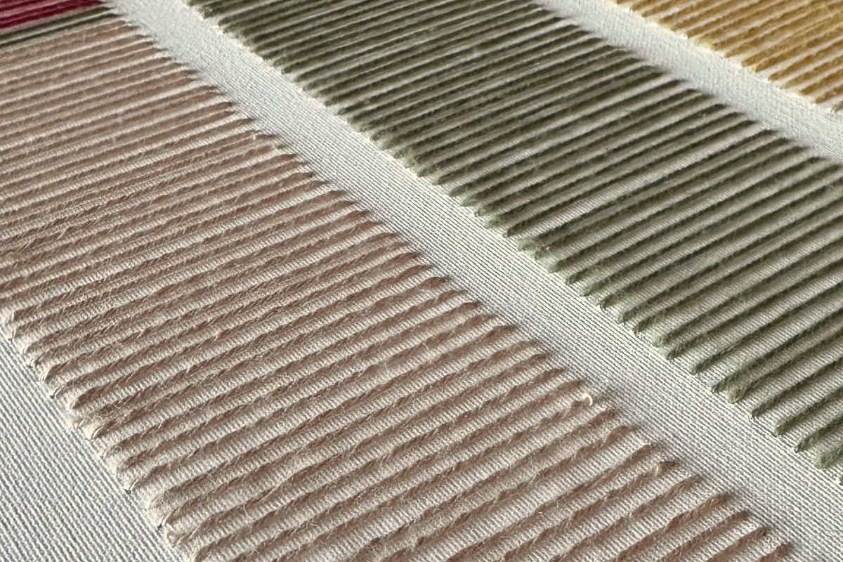 Raking-light detail of Cadence by Nyra Atelier: parallel hand-embroidered wool stitches in blush and olive green, with mustard and red rows beyond, on white canvas
