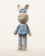 Celeste, handgemachter Häkelhase von Nyra Atelier in einem himmelblauen Rüschenkleid und mit blauer Blumenschleife, mit blau-beige gestreiften Armen und Beinen, Studiofoto auf warmem Elfenbeinhintergrund