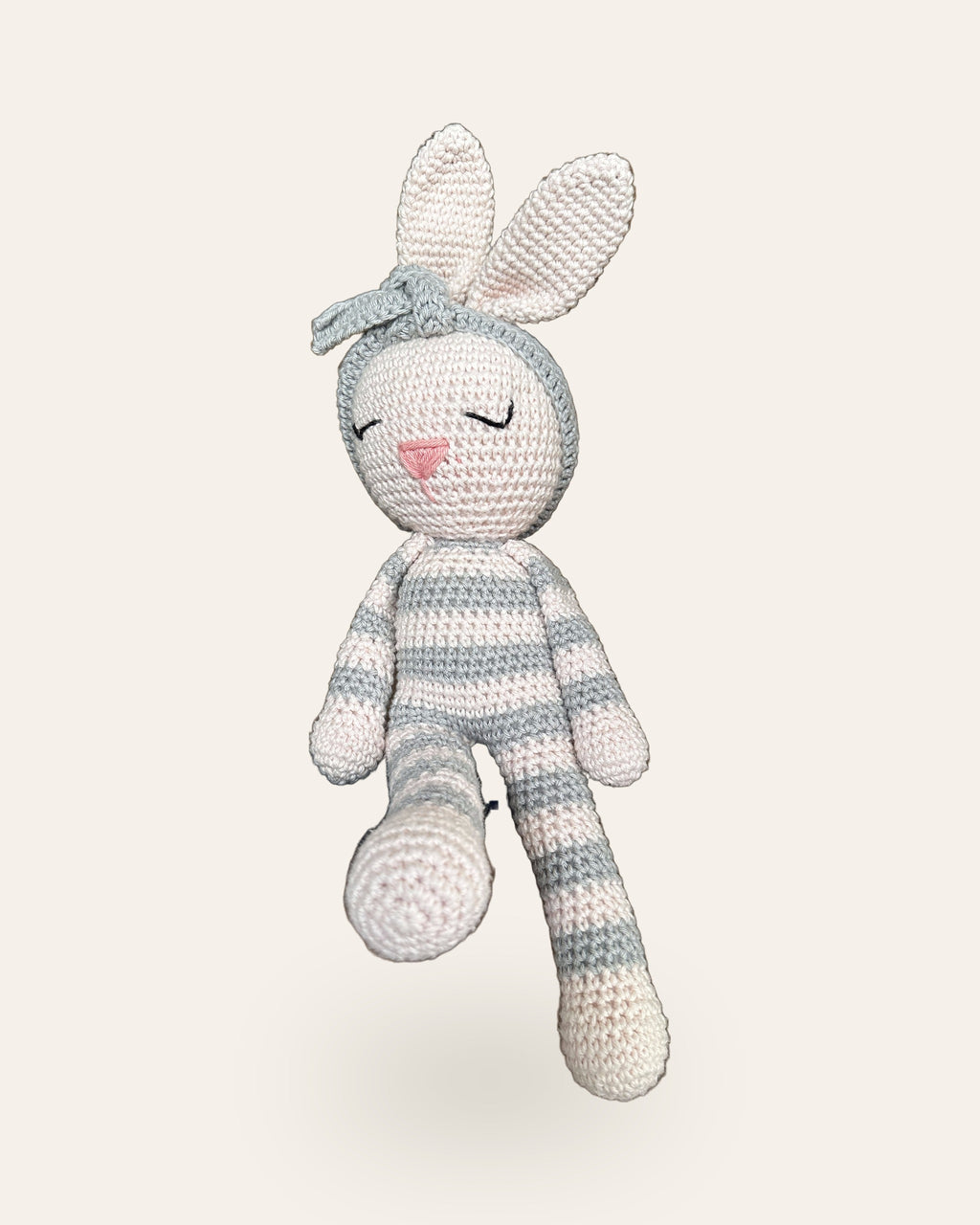 Luna, handmade crochet bunny by Nyra Atelier with grey and cream stripes and her grey flower headband, sitting with her legs dangling. Real photo, cut out on a warm ivory background.