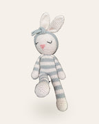 Luna, handmade crochet bunny by Nyra Atelier with grey and cream stripes and her grey flower headband, sitting with her legs dangling. Real photo, cut out on a warm ivory background.