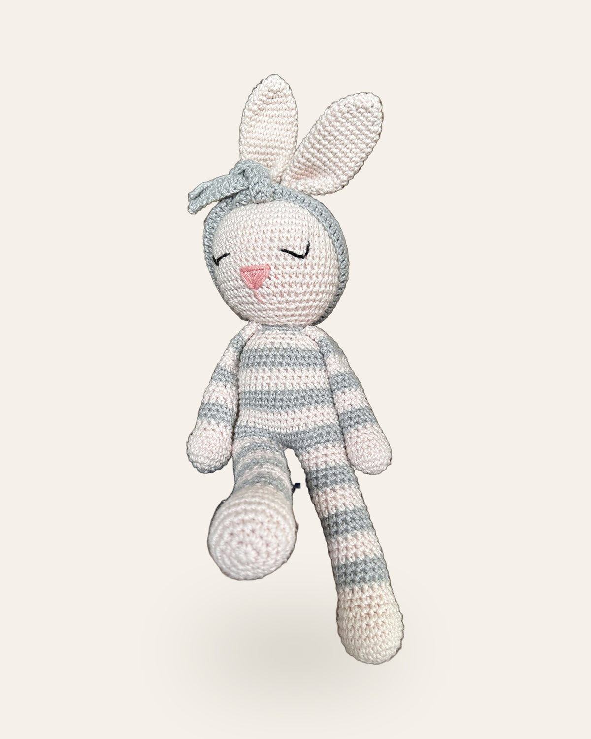 Luna, handmade crochet bunny by Nyra Atelier with grey and cream stripes and her grey flower headband, sitting with her legs dangling. Real photo, cut out on a warm ivory background.