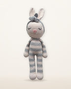 Luna, handmade crochet bunny by Nyra Atelier with grey and cream stripes, pale pink inner ears and a grey flower headband, studio photo on a warm ivory background