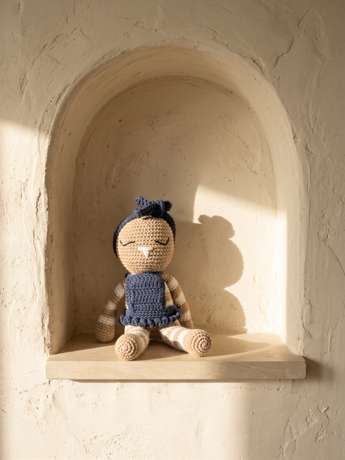 Mia, handmade crochet bunny by Nyra Atelier in her navy blue dress, sitting in a rounded niche of cream lime plaster in the afternoon sun. AI-assisted lifestyle image: the setting is illustrative, the piece is real; please refer to our real product photographs for the actual item's details.