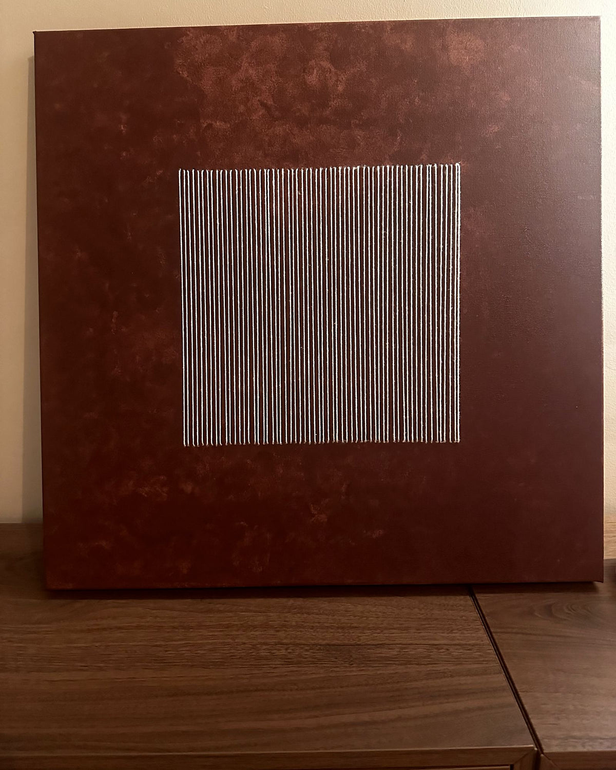 Legato an ein Nussbaum-Sideboard vor einer cremefarbenen Wand gelehnt, das elfenbeinfarbene Fadenquadrat leuchtet auf dem Erdrot