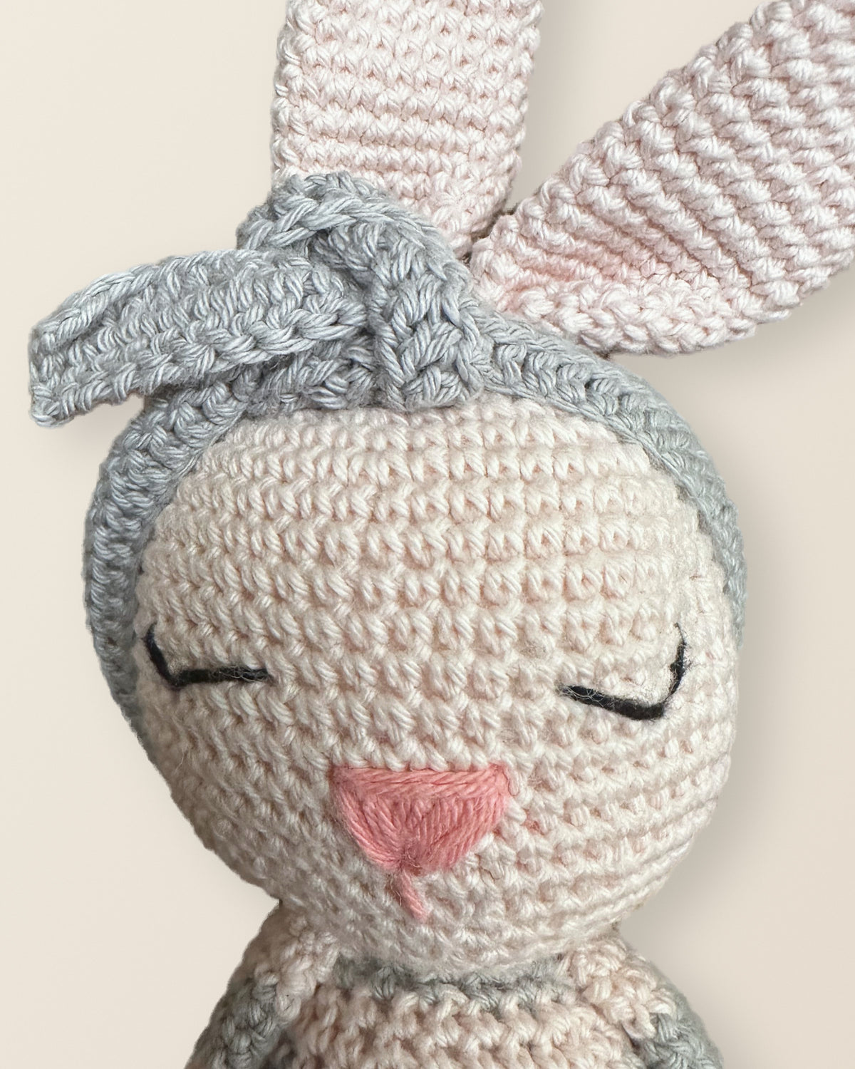 Macro of Luna, a handmade crochet bunny by Nyra Atelier: tight single-crochet stitches in blush cotton, embroidered closed eyes in black thread, a pink satin-stitch nose and a knotted grey crochet headband beneath tall pale-pink ears
