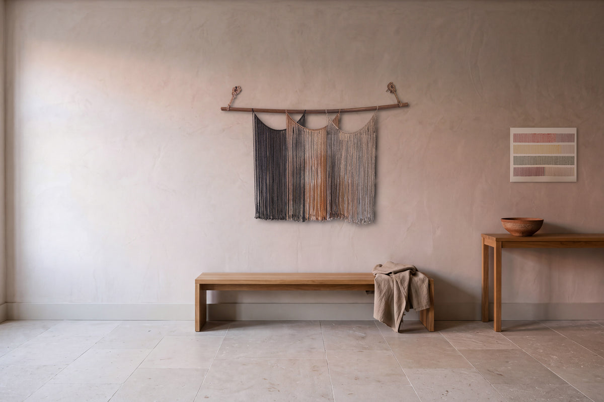 Suspenso Terra macramé wall hanging above a long oak bench, with the Cadence embroidered canvas over an oak console with a terracotta bowl, in a rosy plaster hall with a limestone floor. AI-assisted setting: the room was created with AI and the pieces are real.