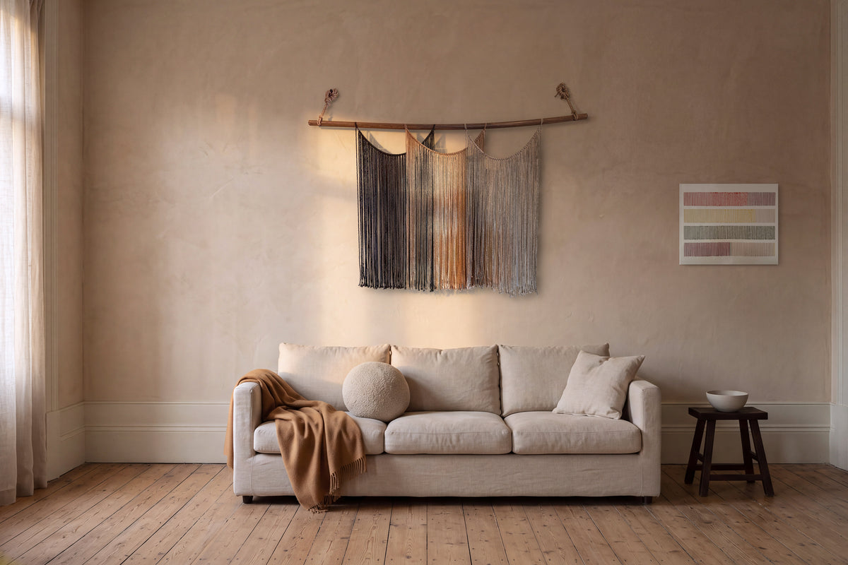 Suspenso Terra, ein 170 cm breiter handgefertigter Makramee-Wandbehang, und Cadence, handbestickte Textilkunst auf Leinwand, von Nyra Atelier in einem Raum mit Putzwänden über einem Leinensofa, mit goldenem Spätnachmittagslicht an der Wand. KI-Szenerie: Der Raum wurde mit KI erstellt; die Stücke sind echte Fotografien.