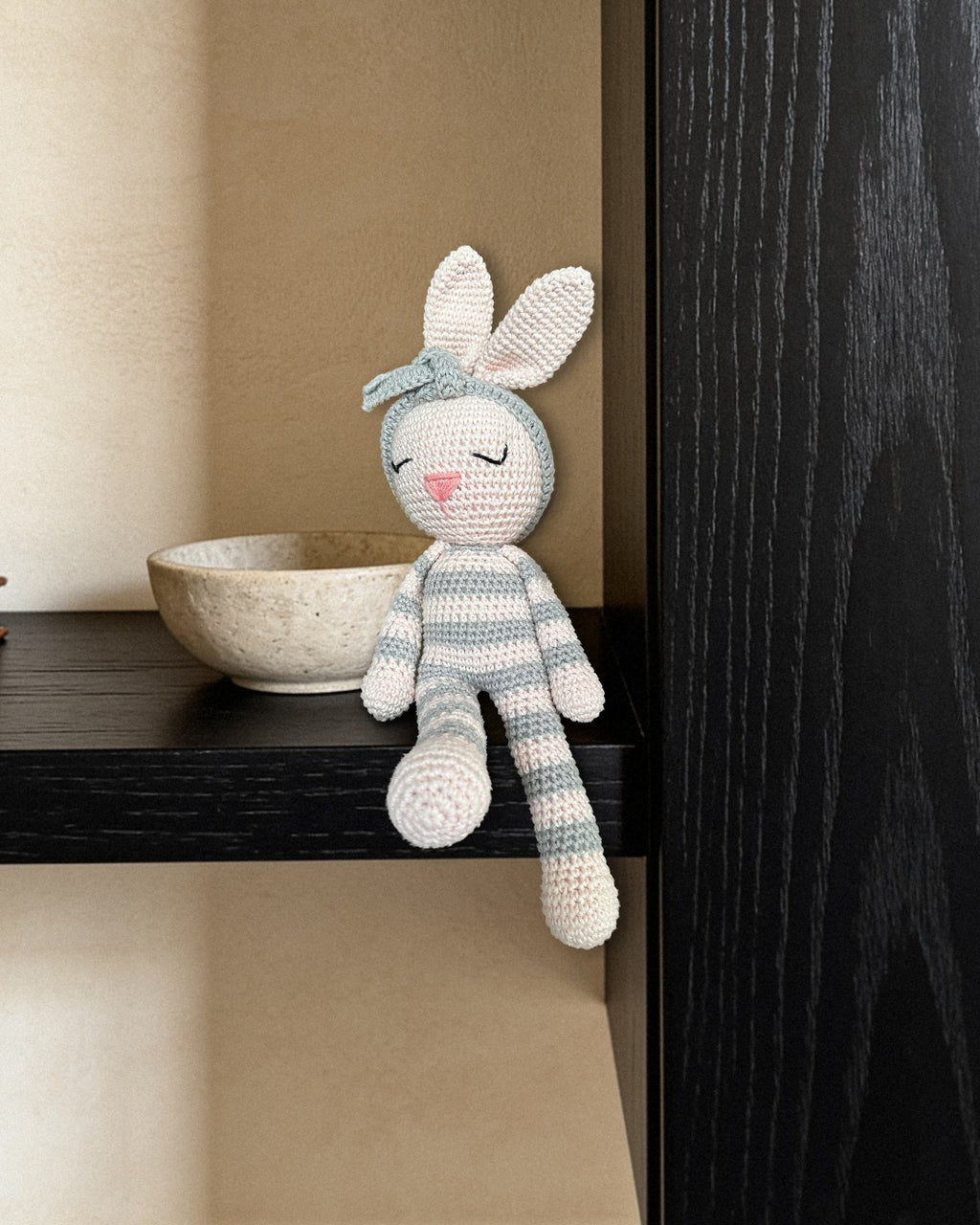 Luna da Nyra Atelier, coelhinha de croché feita à mão, às riscas cinzentas e cremes, com bandolete de flor cinzenta, peça decorativa de coleção sentada na borda de uma prateleira de carvalho tingido quase preto ao lado de uma taça de pedra clara, contra uma parede de estuque de cal cor de areia, em luz suave. Peça real (cenário IA)