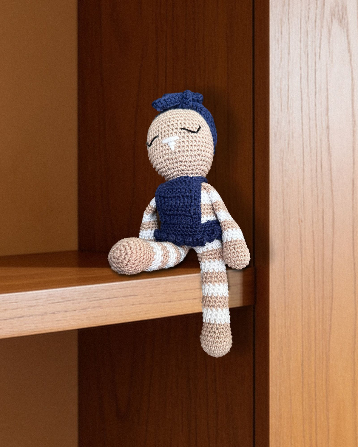 Mia de Nyra Atelier, lapine au crochet faite main, en robe bleu marine à volants et bandeau à nœud, objet décoratif de collection assis au bord d’une étagère en noyer couleur miel, devant un mur d’enduit à la chaux caramel, dans une douce lumière du jour. Pièce réelle (décor IA)