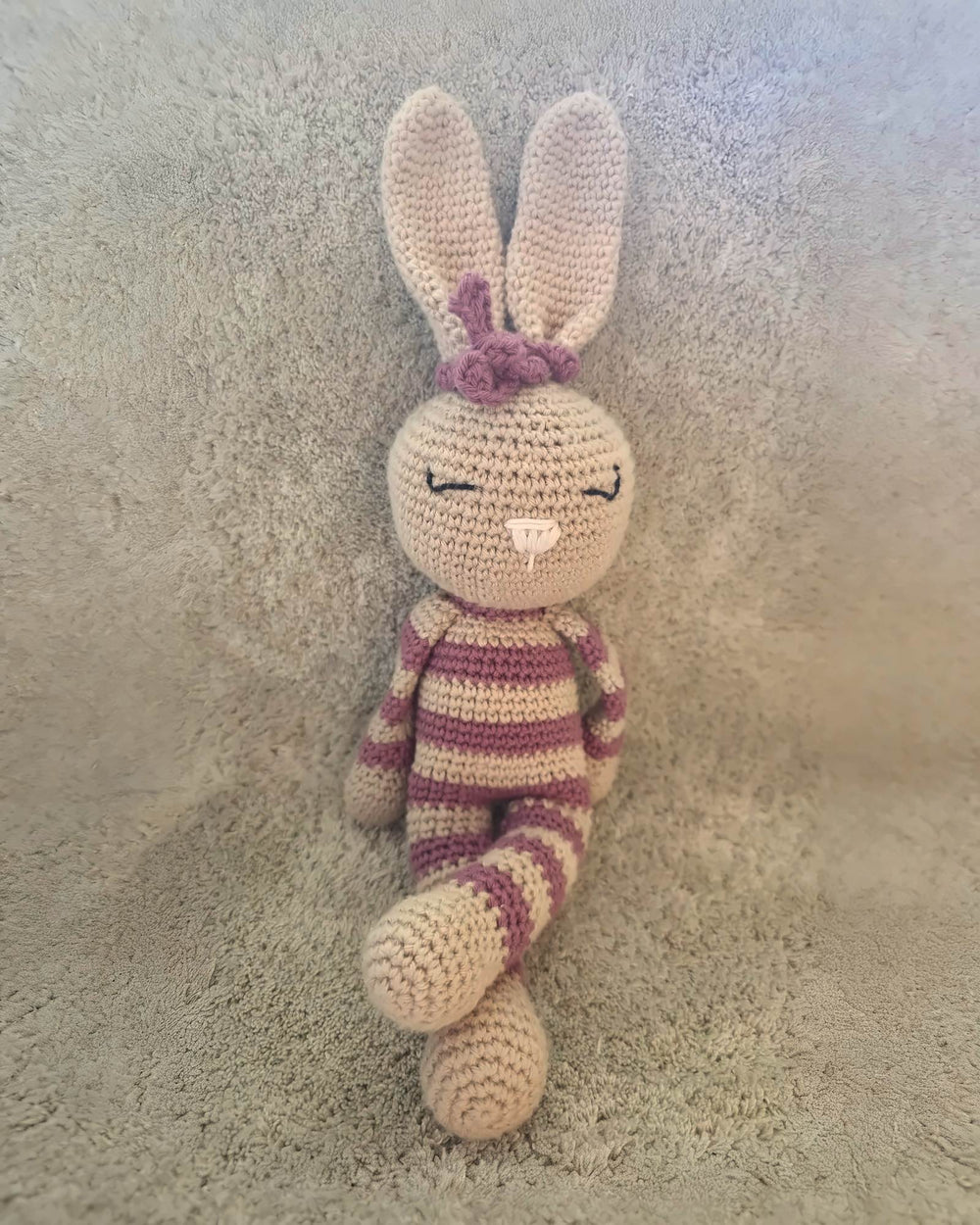 Rosa, lapine au crochet faite main par Nyra Atelier, à rayures roses et beiges, avec un nœud rose et une jambe croisée. Photo réelle ; bords du fond prolongés par IA.