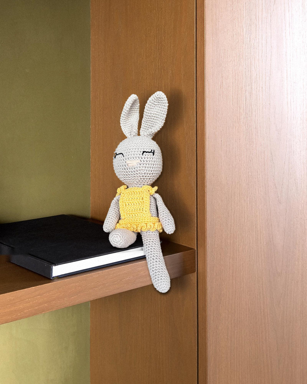 Sol di Nyra Atelier, coniglietta all’uncinetto fatta a mano, con corpo grigio chiaro e abito giallo a balze, oggetto decorativo da collezione seduto all’estremità di una mensola in rovere affumicato accanto a un libro d’arte nero, davanti a una parete in intonaco di calce verde oliva, nella luce morbida del giorno. Pezzo reale (scena IA)