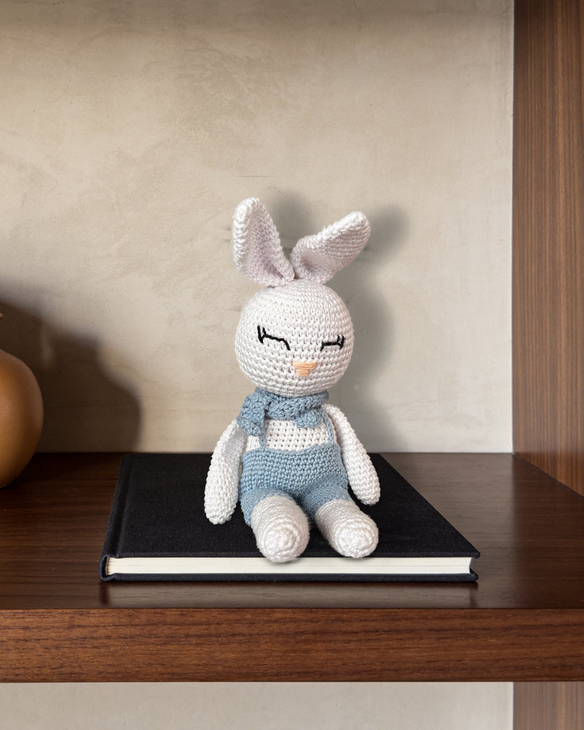 Theo by Nyra Atelier, handmade white crochet bunny in light blue overalls and a neck bow, a decorative collectible sitting on a black linen art book on a dark walnut bookcase shelf, against a stone-grey lime plaster wall in soft daylight. Real piece (AI setting)