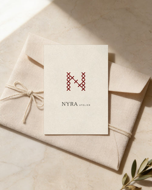 Cartão-oferta da Nyra Atelier em papel de algodão marfim, com um N vermelho em ponto de cruz e letras finas, sobre um envelope de linho atado com fio de algodão, em travertino claro. (cenário IA)