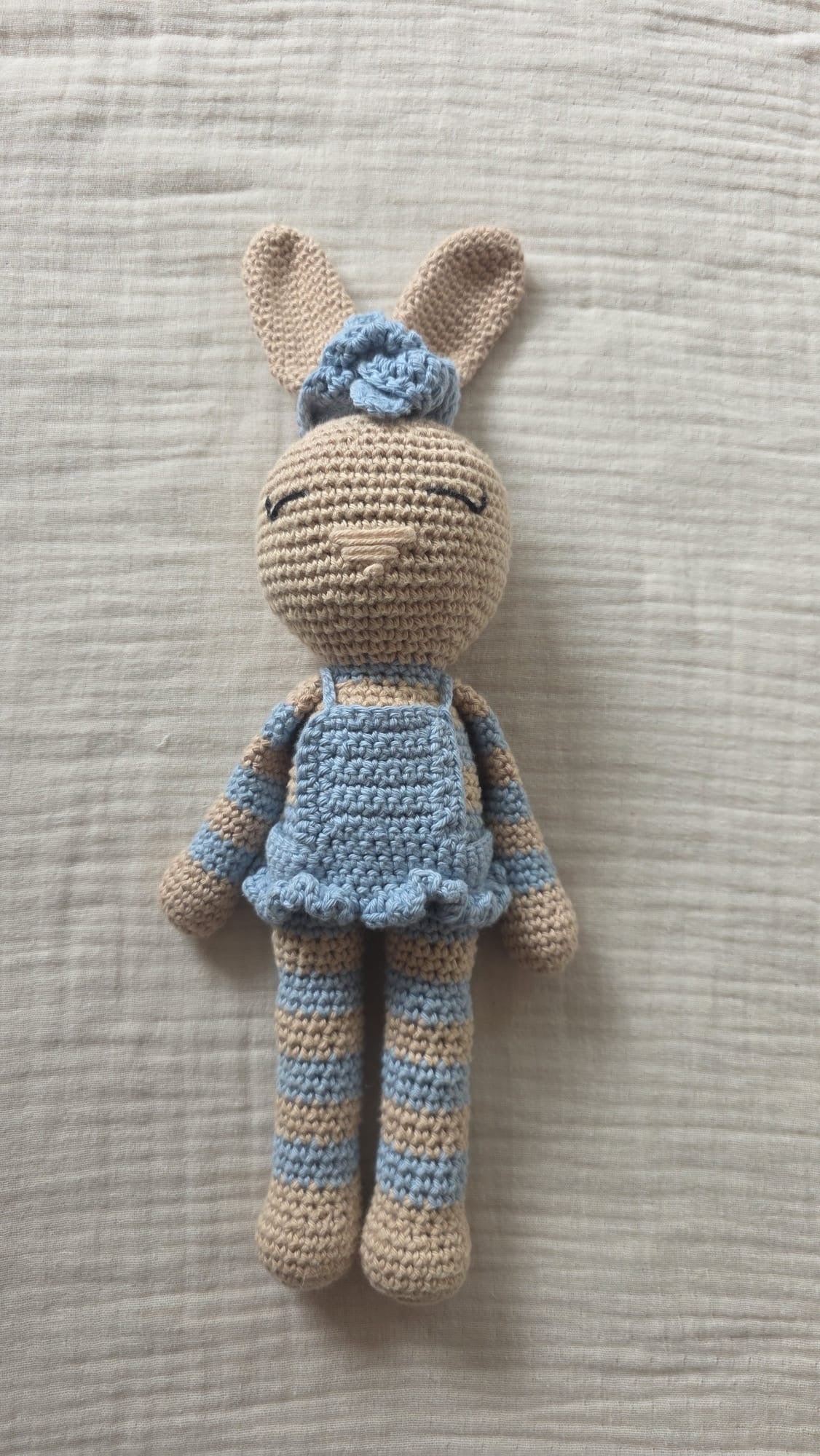 Celeste, handgemachter Häkelhase in einem himmelblauen Rüschenkleid und mit blauer Blumenschleife, mit blau-beige gestreiften Armen und Beinen, stehend auf Leinen