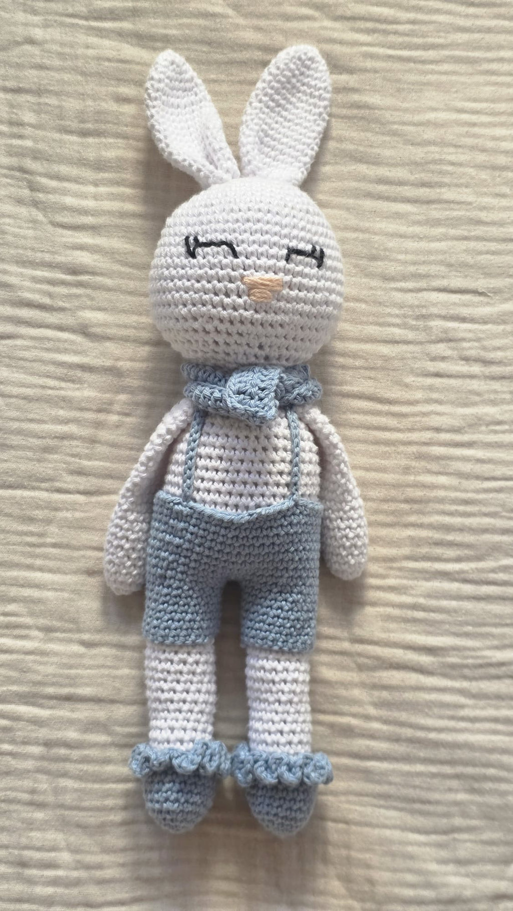 Theo, handmade white crochet bunny with long ears, light blue overalls and a neck bow, standing on linen