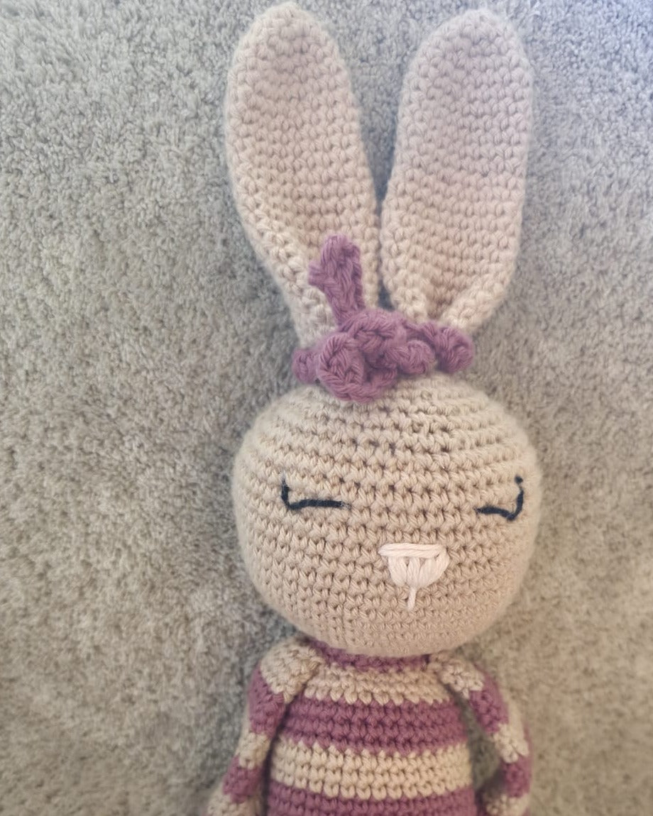 Close-up of Rosa, handmade crochet bunny: tan ears, a pink crochet bow, embroidered sleeping eyes and pink-and-beige stripes
