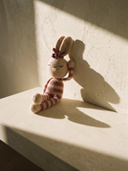 Rosa, handmade crochet bunny by Nyra Atelier with pink and cream stripes, leaning against a lime-plaster wall on a block of limestone, with a long afternoon shadow. AI-assisted lifestyle image: the setting is illustrative, the piece is real; please refer to our real product photographs for the actual item's details.