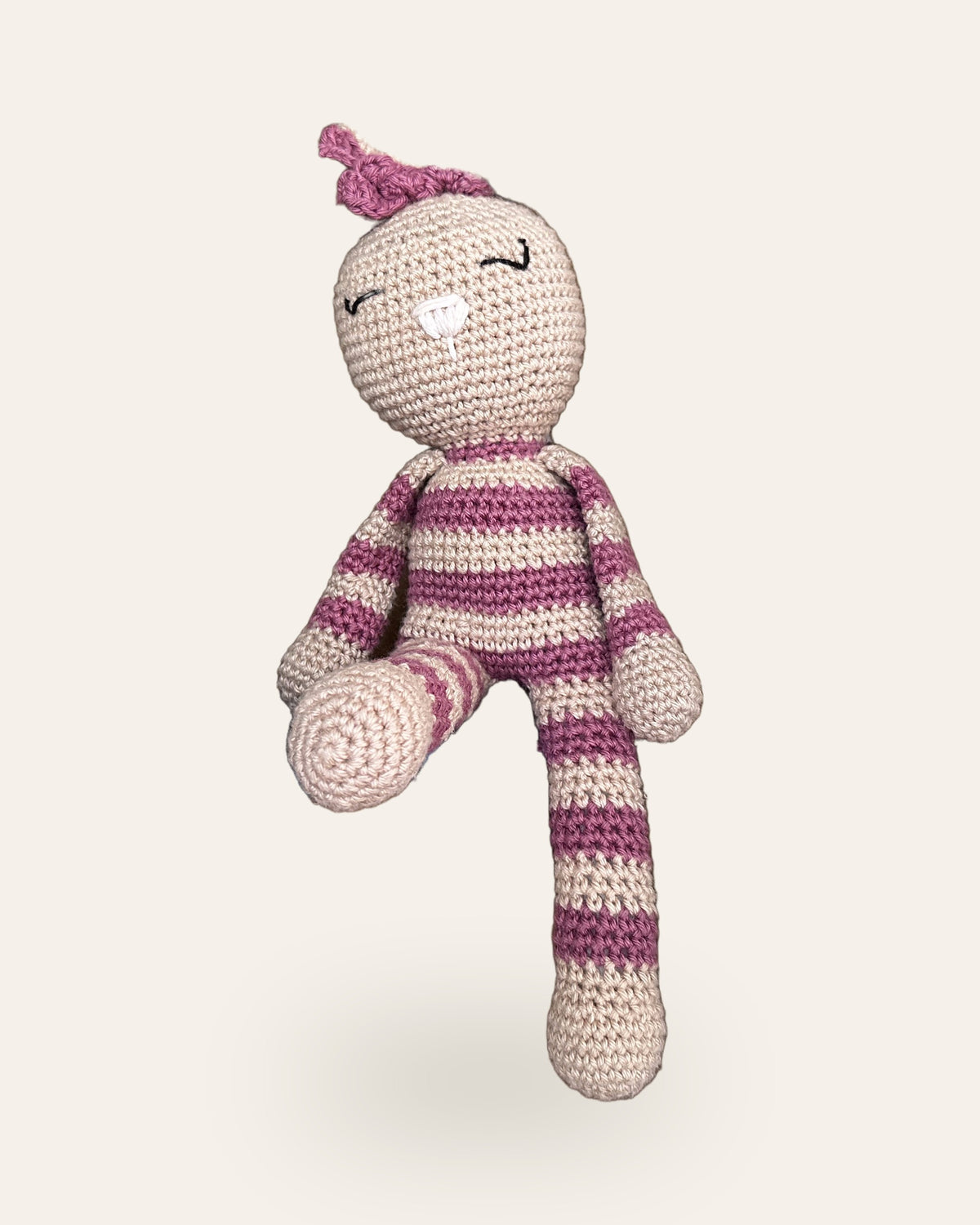 Rosa, handmade crochet bunny by Nyra Atelier with pink and beige stripes and a pink bow, sitting with one leg tucked in. Real photo, cut out on a warm ivory background.