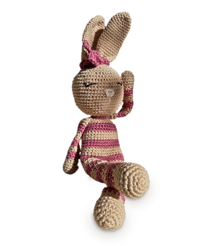Rosa, a handmade crochet bunny by Nyra Atelier with tan ears, a raspberry pink bow and raspberry-and-beige stripes, one paw raised to her ear and one leg crossed, seated