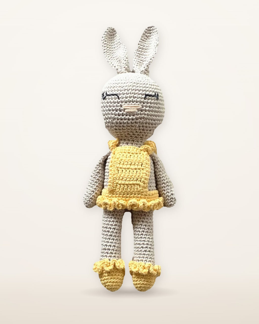 Sol, handmade crochet bunny by Nyra Atelier with a pale grey body, long ears, a yellow ruffled dress and yellow ruffled shoes, studio photo on a warm ivory background