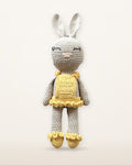 Sol, handmade crochet bunny by Nyra Atelier with a pale grey body, long ears, a yellow ruffled dress and yellow ruffled shoes, studio photo on a warm ivory background