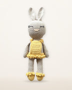 Sol, handmade crochet bunny by Nyra Atelier with a pale grey body, long ears, a yellow ruffled dress and yellow ruffled shoes, studio photo on a warm ivory background