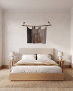 Suspenso Terra macramé wall hanging above a low oak bed with linen bedding and two ceramic lamps. AI setting: the room was created with AI and the piece is real.
