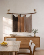 Suspenso Terra macramé wall hanging above a low oak sideboard in a calm dining room, with a travertine table and a bowl of lemons in the foreground. AI setting: the room was created with AI and the piece is real.