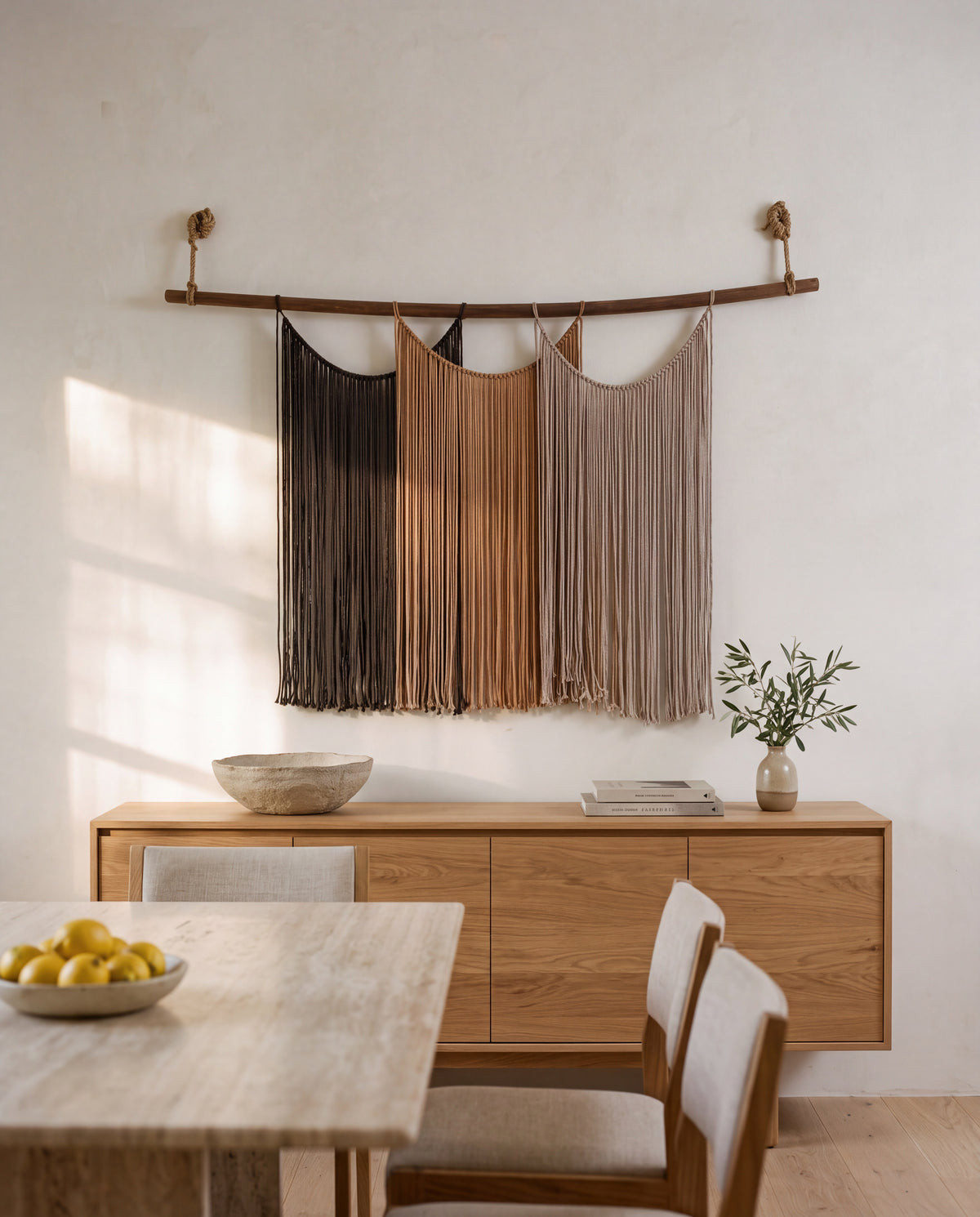 Suspenso Terra macramé wall hanging above a low oak sideboard in a calm dining room, with a travertine table and a bowl of lemons in the foreground. AI setting: the room was created with AI and the piece is real.