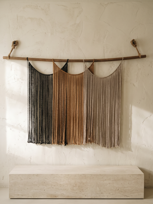 Suspenso Terra, large handmade macramé wall hanging by Nyra Atelier, on a cream lime-plaster wall above a limestone plinth in soft morning light. AI setting: the room was created with AI and the piece is real.