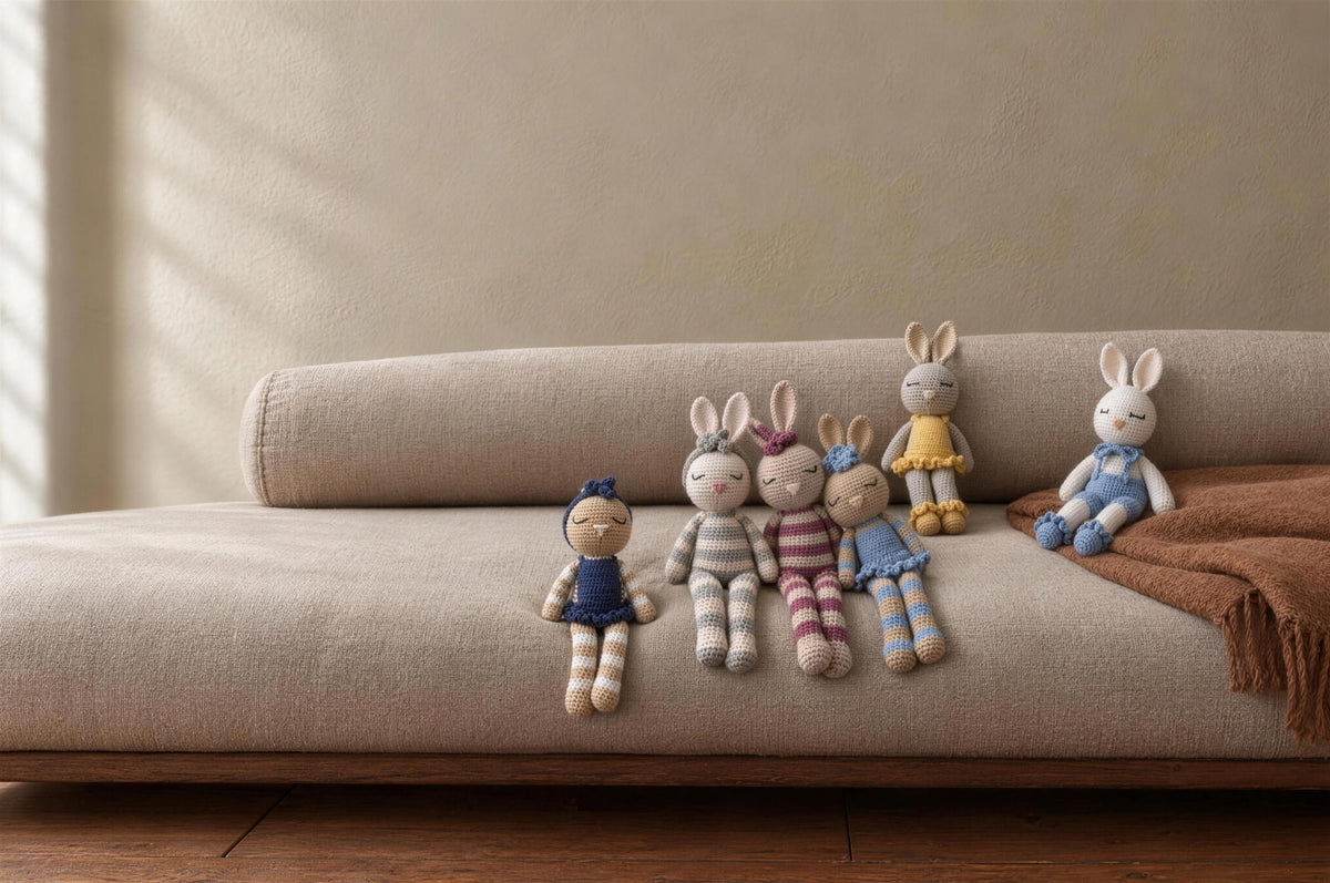 The Nyra Bunny Family, six handmade crochet bunnies by Nyra Atelier: Mia in a navy dress, Luna in grey stripes, Rosa in raspberry stripes, Celeste in a sky-blue dress, Sol in a yellow ruffled dress and Theo in blue overalls, seated on an oatmeal linen daybed against a plaster wall in soft north light. AI-assisted setting, the pieces are real.