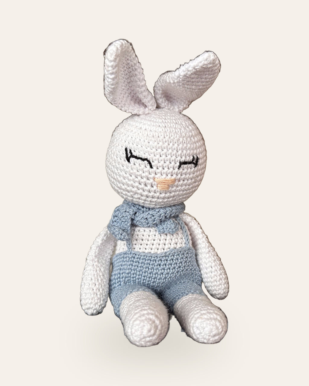 Theo, handmade white crochet bunny by Nyra Atelier, sitting with his legs stretched out, in light blue overalls and a neck bow. Real photo, cut out on a warm ivory background.