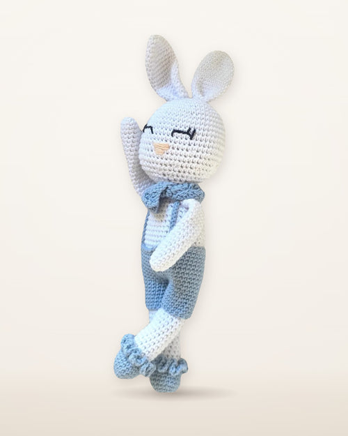 Theo, coniglietto bianco all'uncinetto fatto a mano di Nyra Atelier con orecchie lunghe, salopette azzurra, fiocco al collo e calzini con balze, foto di studio su uno sfondo avorio caldo