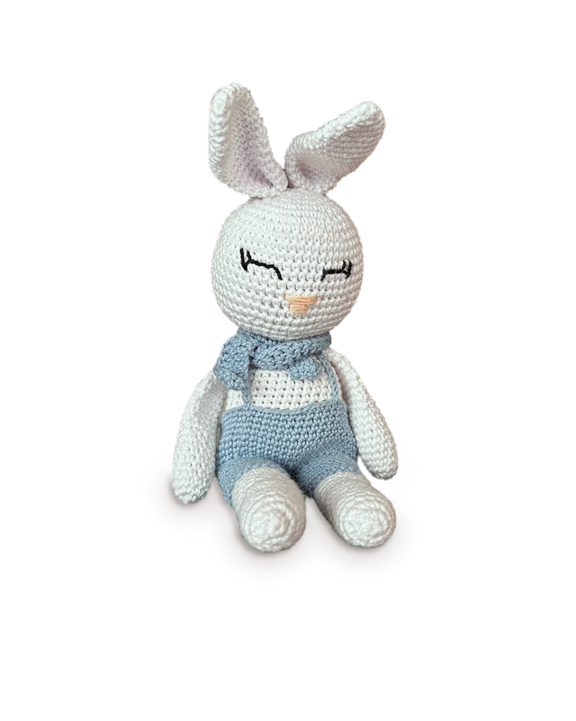 Theo, a handmade white crochet bunny by Nyra Atelier in light blue crochet overalls with a knotted blue neck scarf and long upright ears, seated